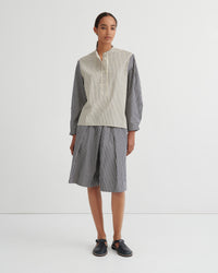 A model stands against a plain white studio background wearing a matching striped two-piece set consisting of a button up shirt and pleated shorts. The shirt has a cream and white vertical stripe pattern on the front and solid dark gray sleeves, while the shorts are entirely dark gray with thin white vertical stripes.