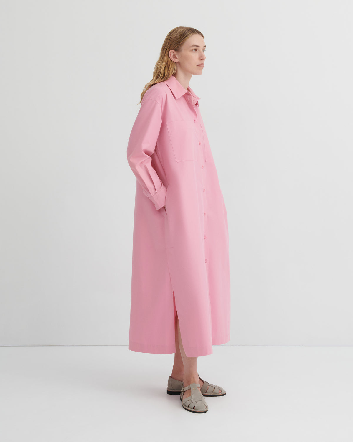A woman modeling a long, light pink button-up shirt dress with a relaxed, oversized fit, shown in profile against a clean white studio background.
