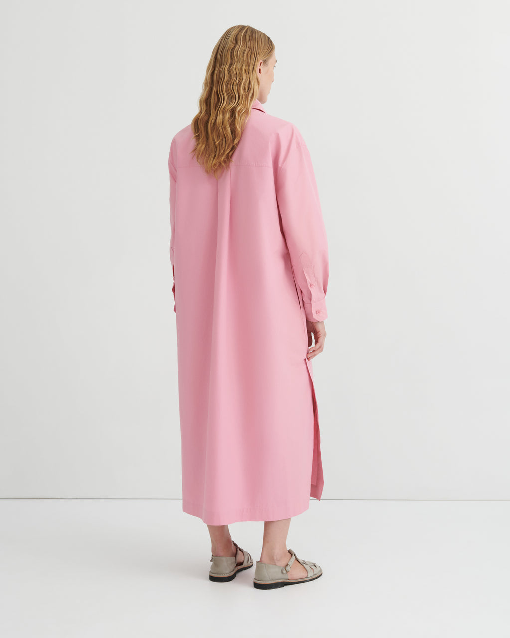 A woman viewed from behind wearing a long pink shirt dress with a collar and side slit, paired with beige sandals, against a plain white studio background.