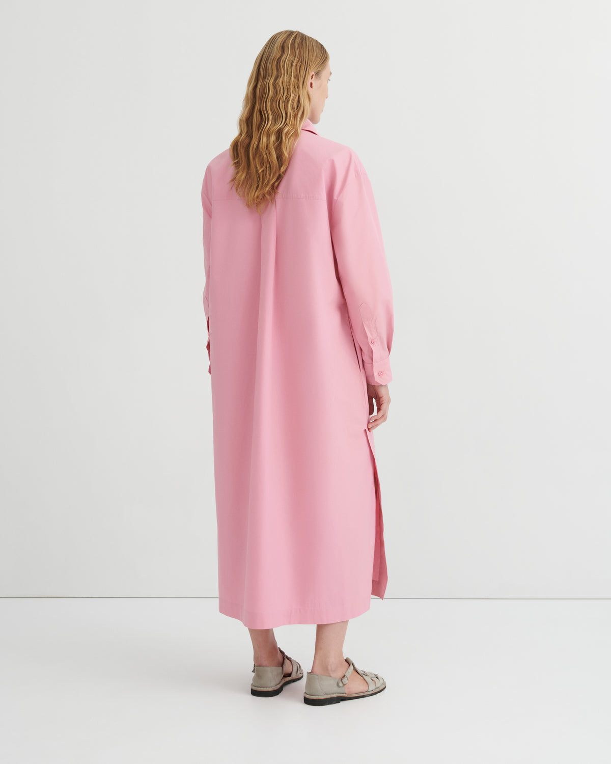 A woman viewed from behind wearing a long pink shirt dress with a collar and side slit, paired with beige sandals, against a plain white studio background.
