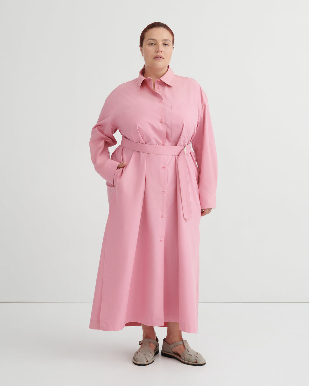 A woman modeling a pink long-sleeve shirt dress with a matching waist tie belt, standing against a plain white studio background.