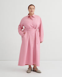 A woman modeling a pink long-sleeve shirt dress with a matching waist tie belt, standing against a plain white studio background.