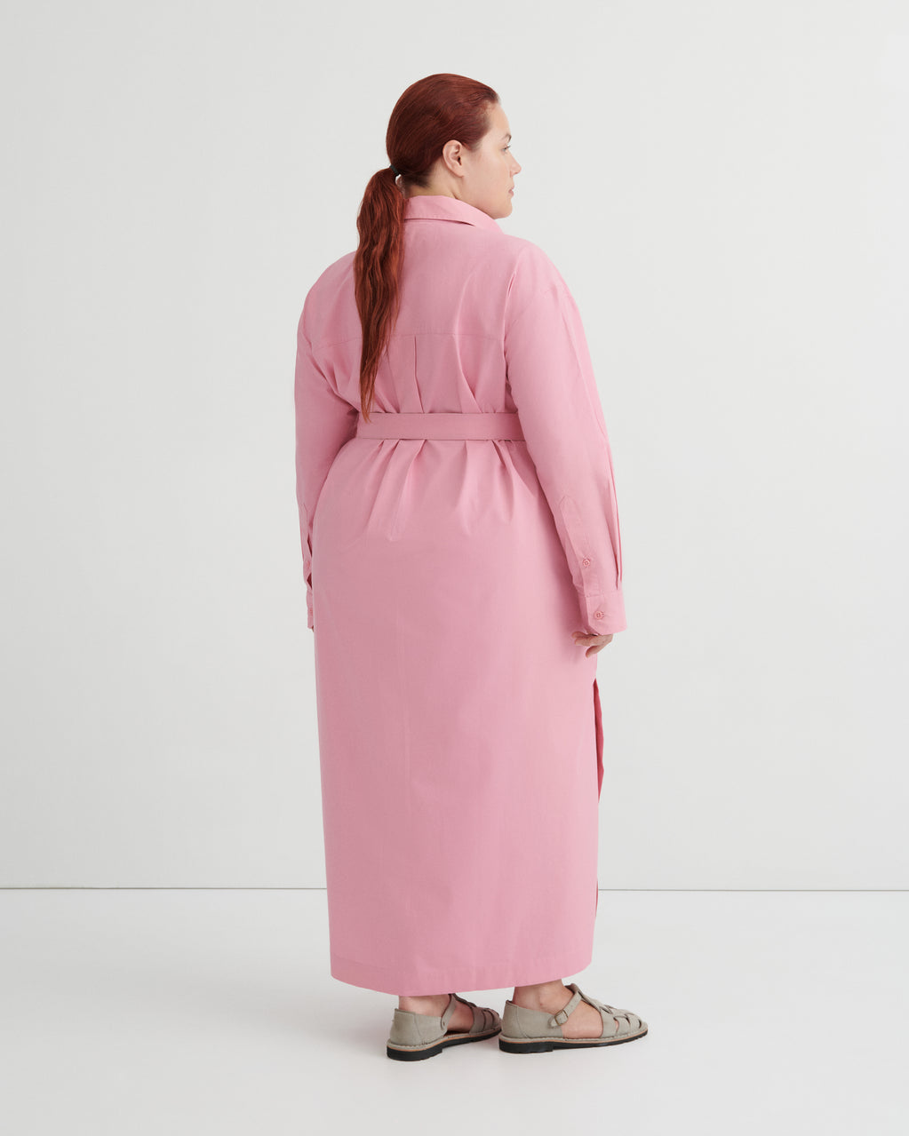 A woman with red hair tied back in a ponytail is shown from the back wearing a long pink belted coat or dress over a dark dress, paired with brown sandals, against a plain white studio background.