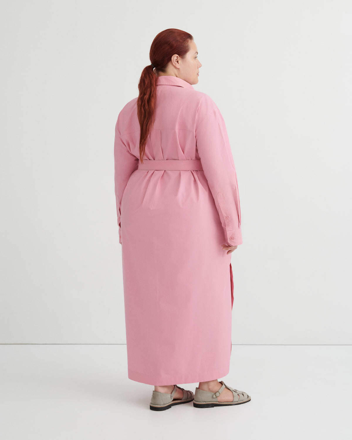 A woman with red hair tied back in a ponytail is shown from the back wearing a long pink belted coat or dress over a dark dress, paired with brown sandals, against a plain white studio background.
