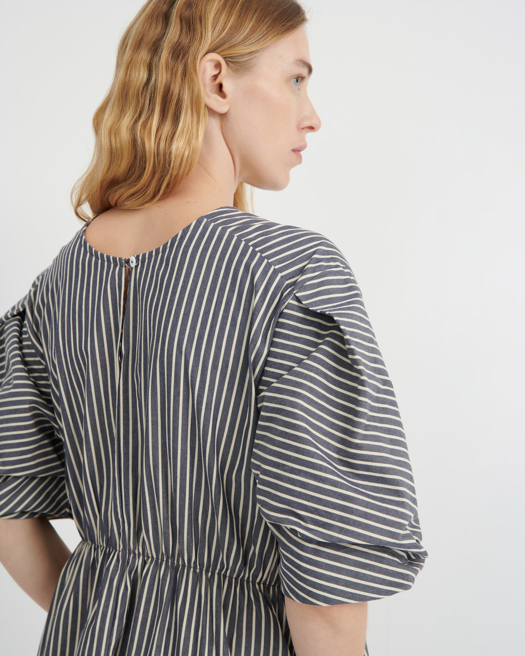 A woman shown from the back wearing a dark gray and white vertically striped dress with puff sleeves and a gathered waist, photographed against a clean white studio background.