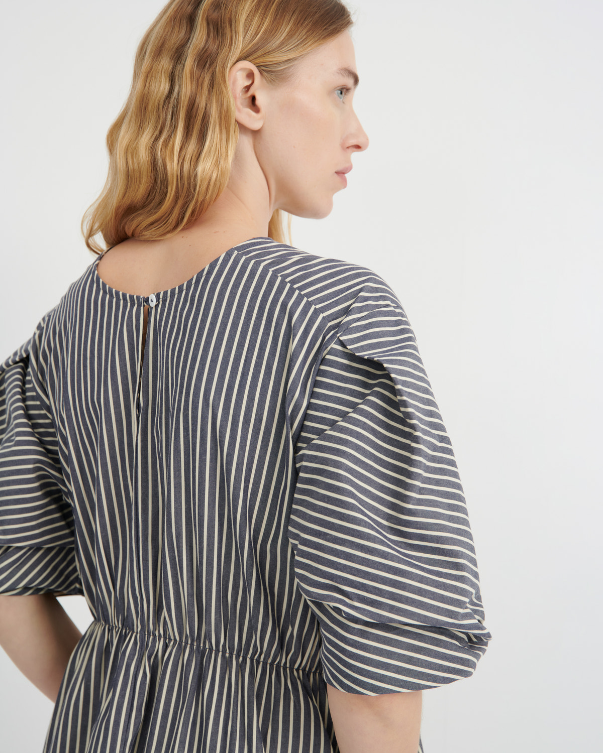 A woman shown from the back wearing a dark gray and white vertically striped dress with puff sleeves and a gathered waist, photographed against a clean white studio background.