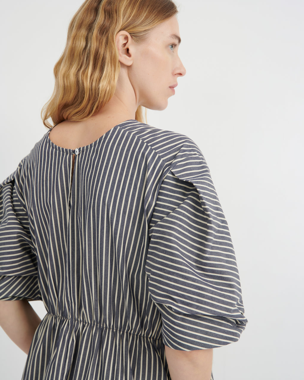 A woman shown from the back wearing a dark gray and white vertically striped dress with puff sleeves and a gathered waist, photographed against a clean white studio background.