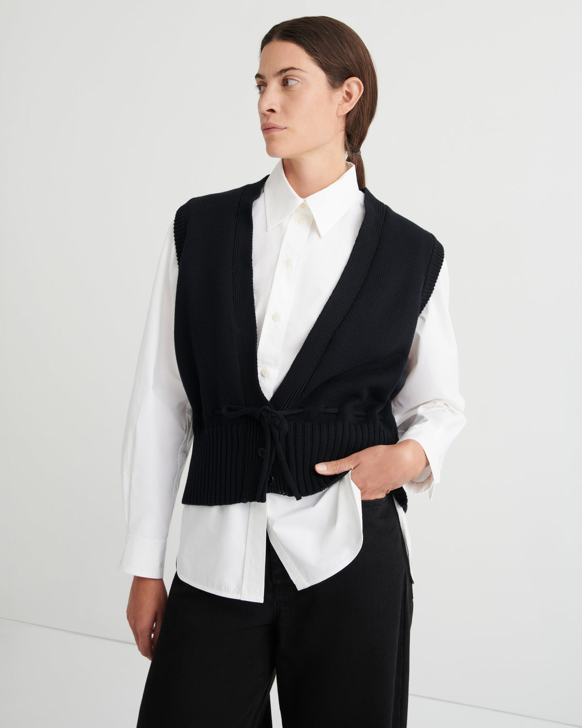 A model wearing a black knit wrap vest over a white collared shirt and black trousers, photographed against a clean white studio background.
