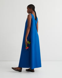 A woman wearing a sleeveless, floor-length royal blue dress, shown from the back and side against a plain light gray studio background.