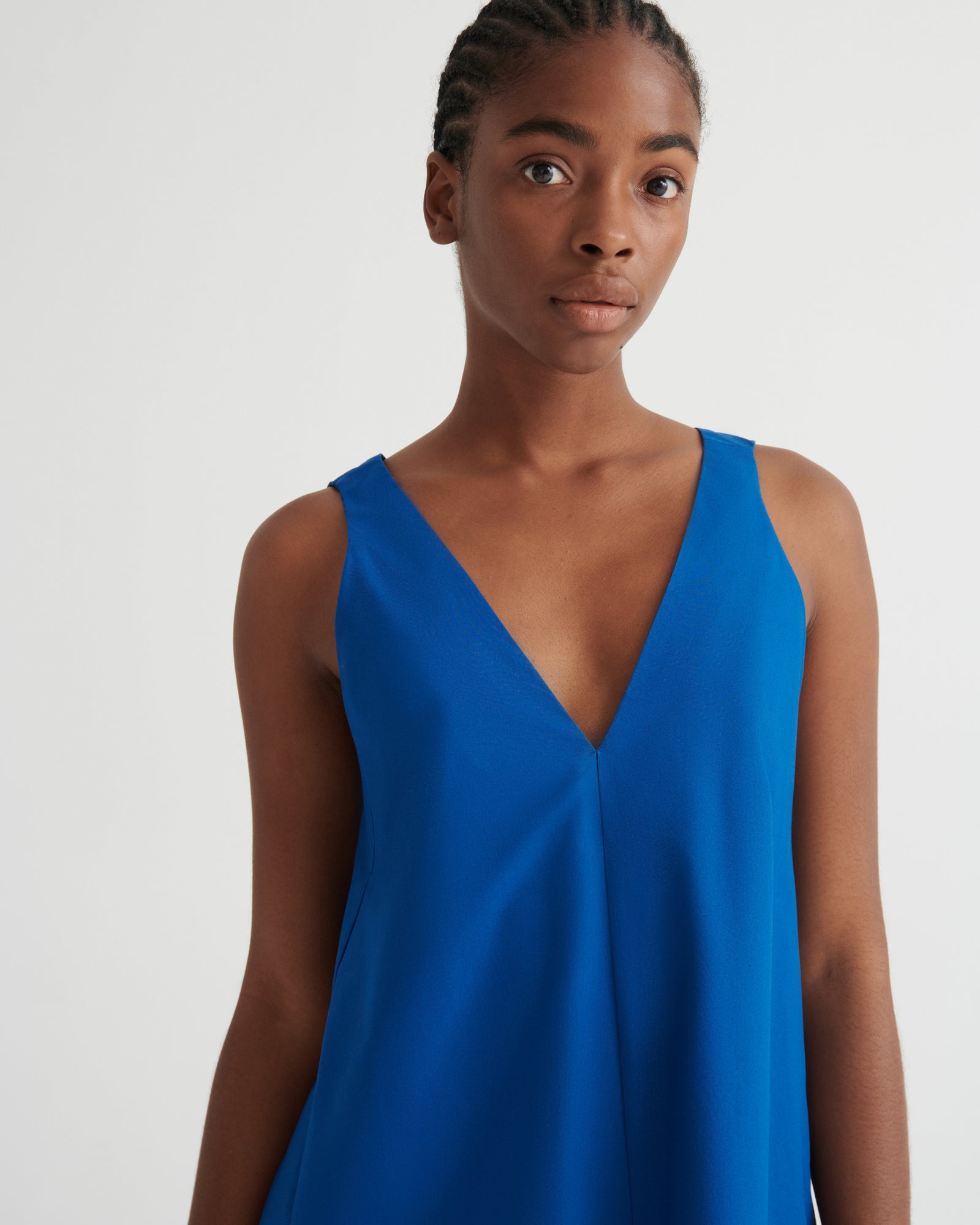 A woman wearing a sleeveless, V-neck blue dress against a plain light gray studio background.