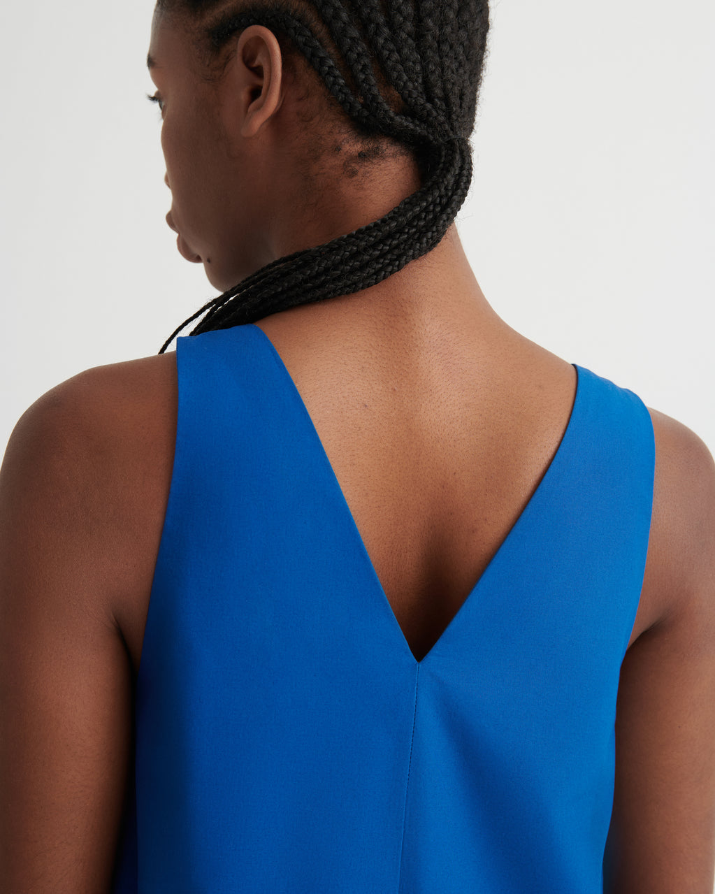 Close-up back view of a woman wearing a bright blue sleeveless dress with a deep V-neckline, against a plain light gray background.