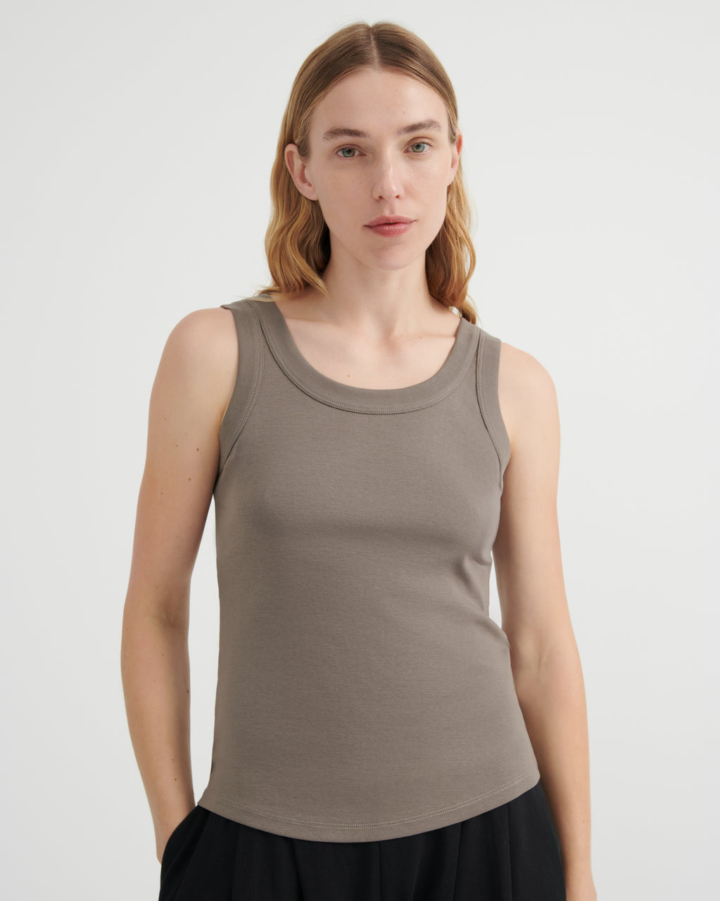 A woman modeling a sleeveless scoop-neck tank top in a muted taupe-brown color, paired with a black skirt.