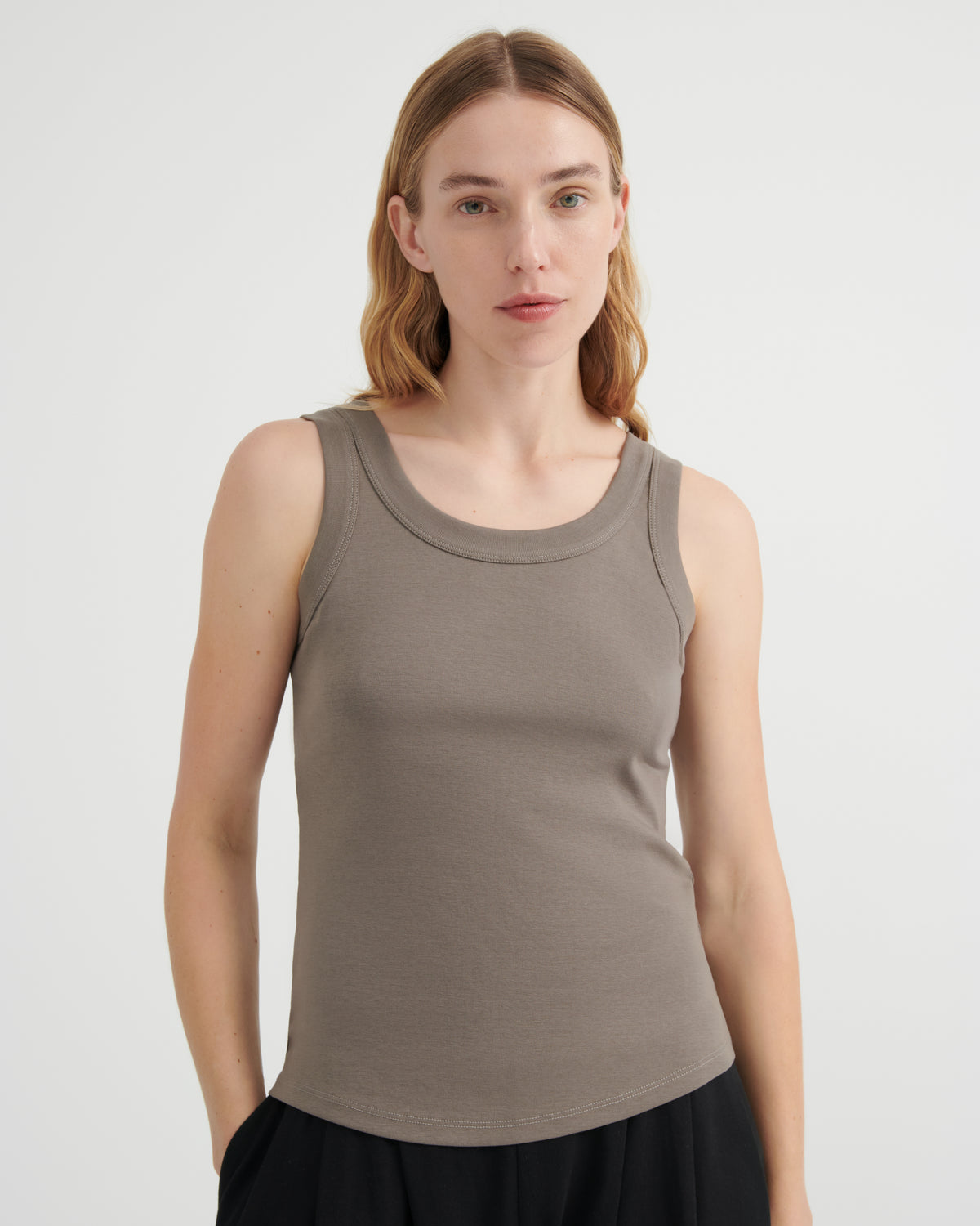 A woman modeling a sleeveless scoop-neck tank top in a muted taupe-brown color, paired with a black skirt.