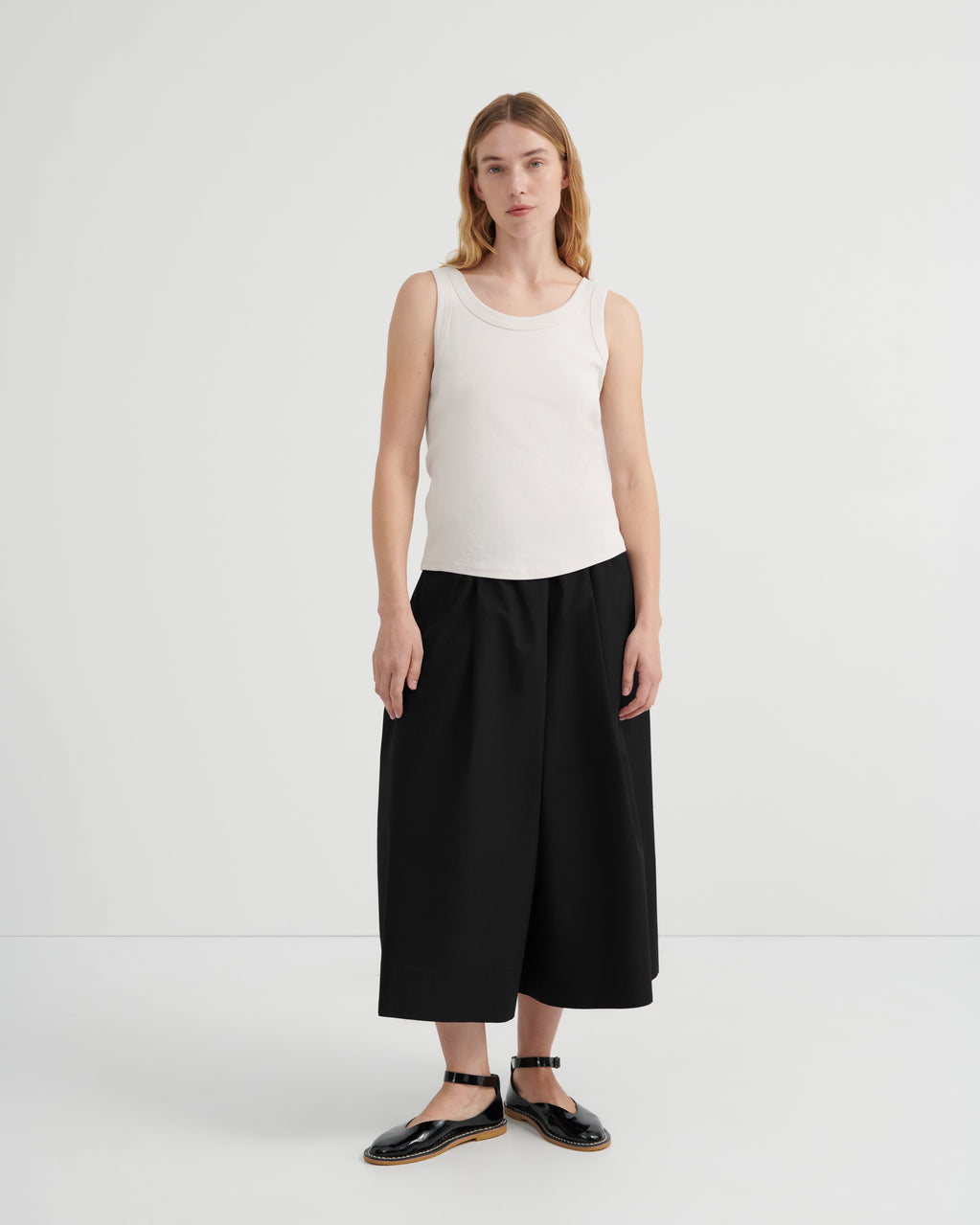 A woman wearing a sleeveless white ribbed tank top, loose black wide-leg cropped pants, and black flat sandals.