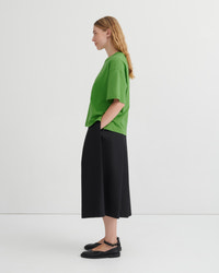A woman stands in profile wearing an oversized bright green short-sleeve top, black wide-leg cropped pants, and black Mary Jane flats against a clean white studio background.