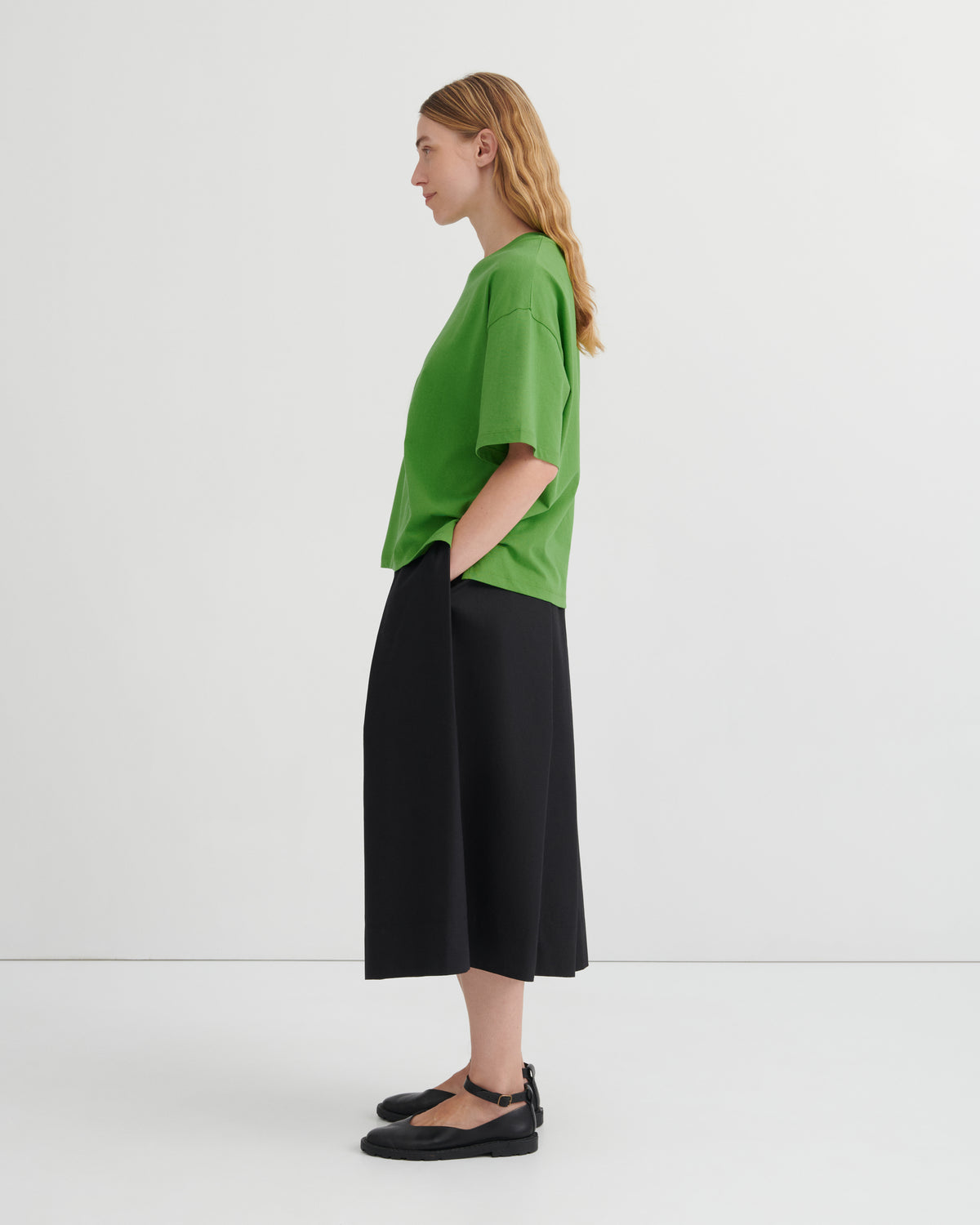 A woman stands in profile wearing an oversized bright green short-sleeve top, black wide-leg cropped pants, and black Mary Jane flats against a clean white studio background.