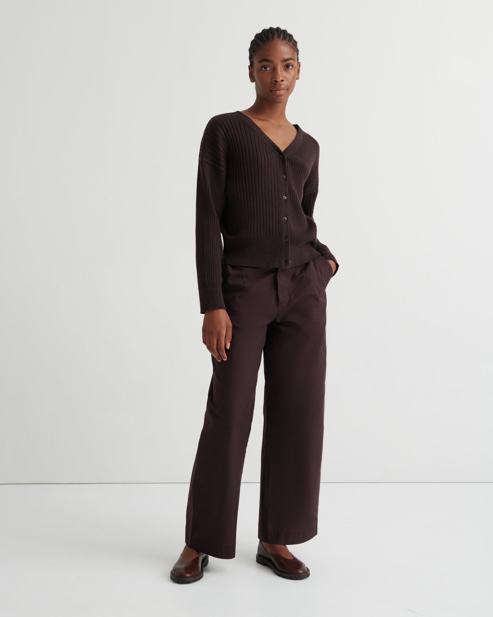 A model stands against a plain light gray studio background wearing a dark brown ribbed knit cardigan with a V-neck and buttons, matching wide-leg trousers, and dark brown loafers.