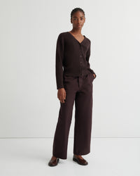 A model stands against a plain light gray studio background wearing a dark brown ribbed knit cardigan with a V-neck and buttons, matching wide-leg trousers, and dark brown loafers.