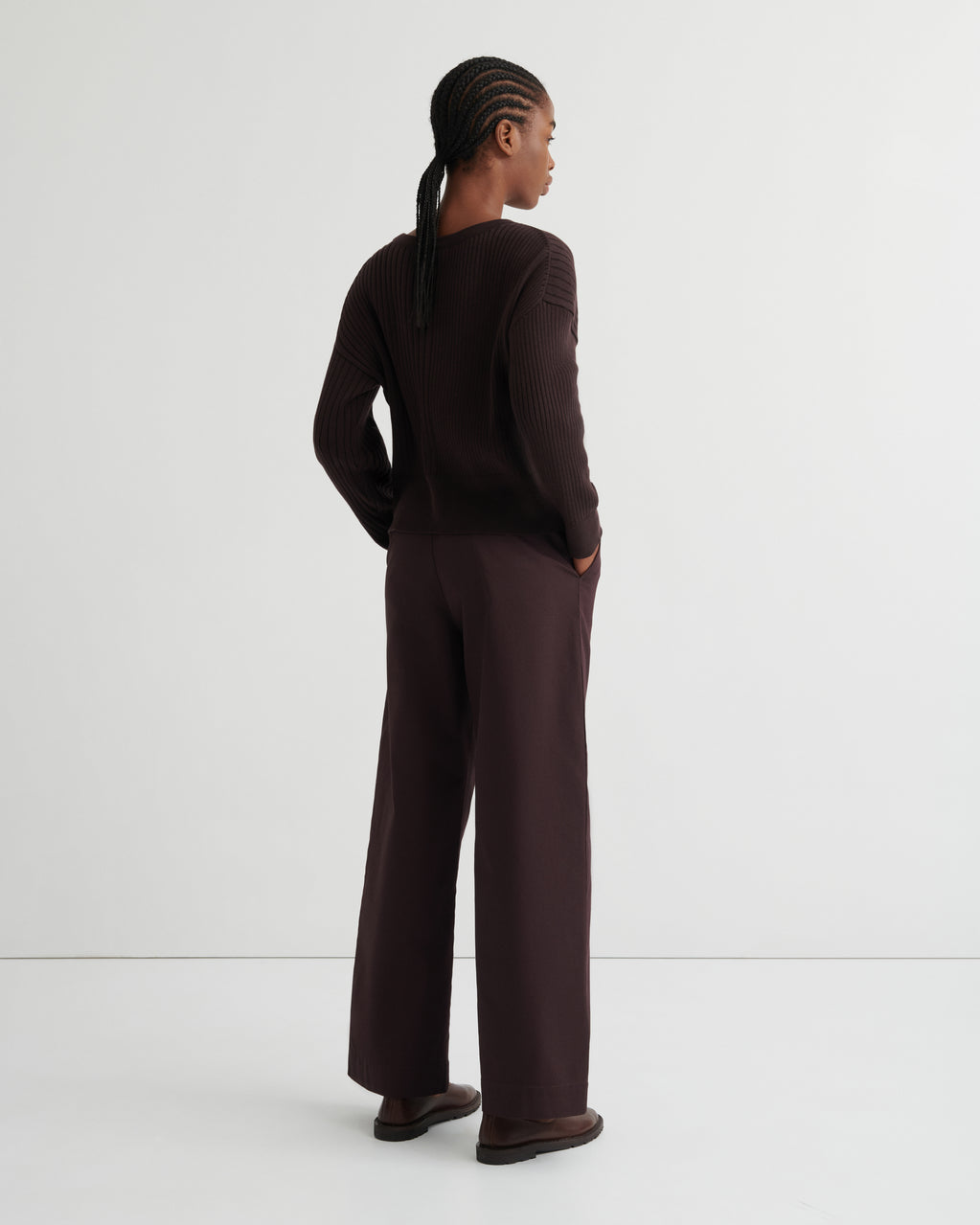 A model is shown from the back wearing a dark brown ribbed knit long-sleeve top and matching wide-leg trousers, standing against a clean white studio background.