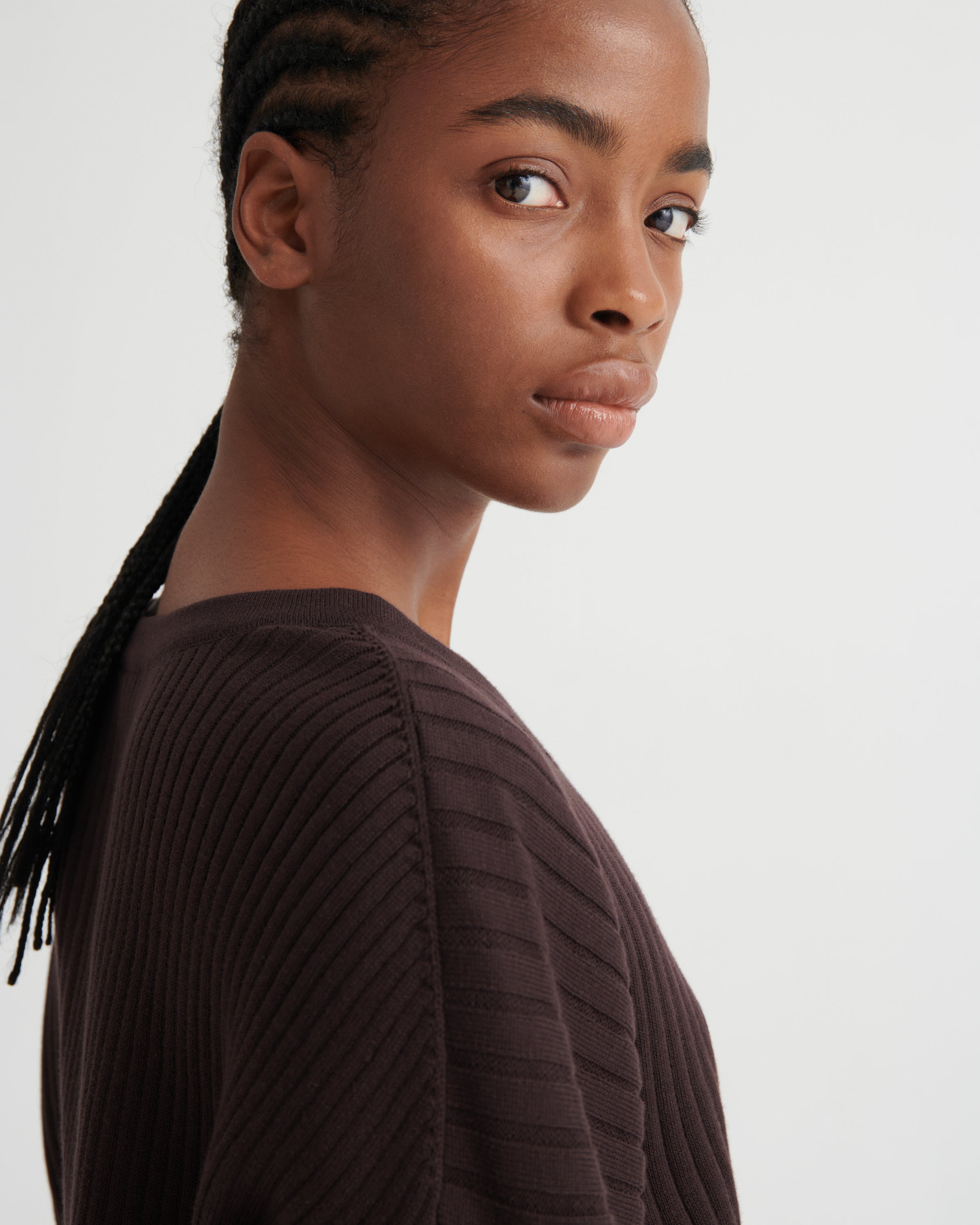 A close-up side profile of a model wearing a dark brown ribbed knit top against a light gray studio background.