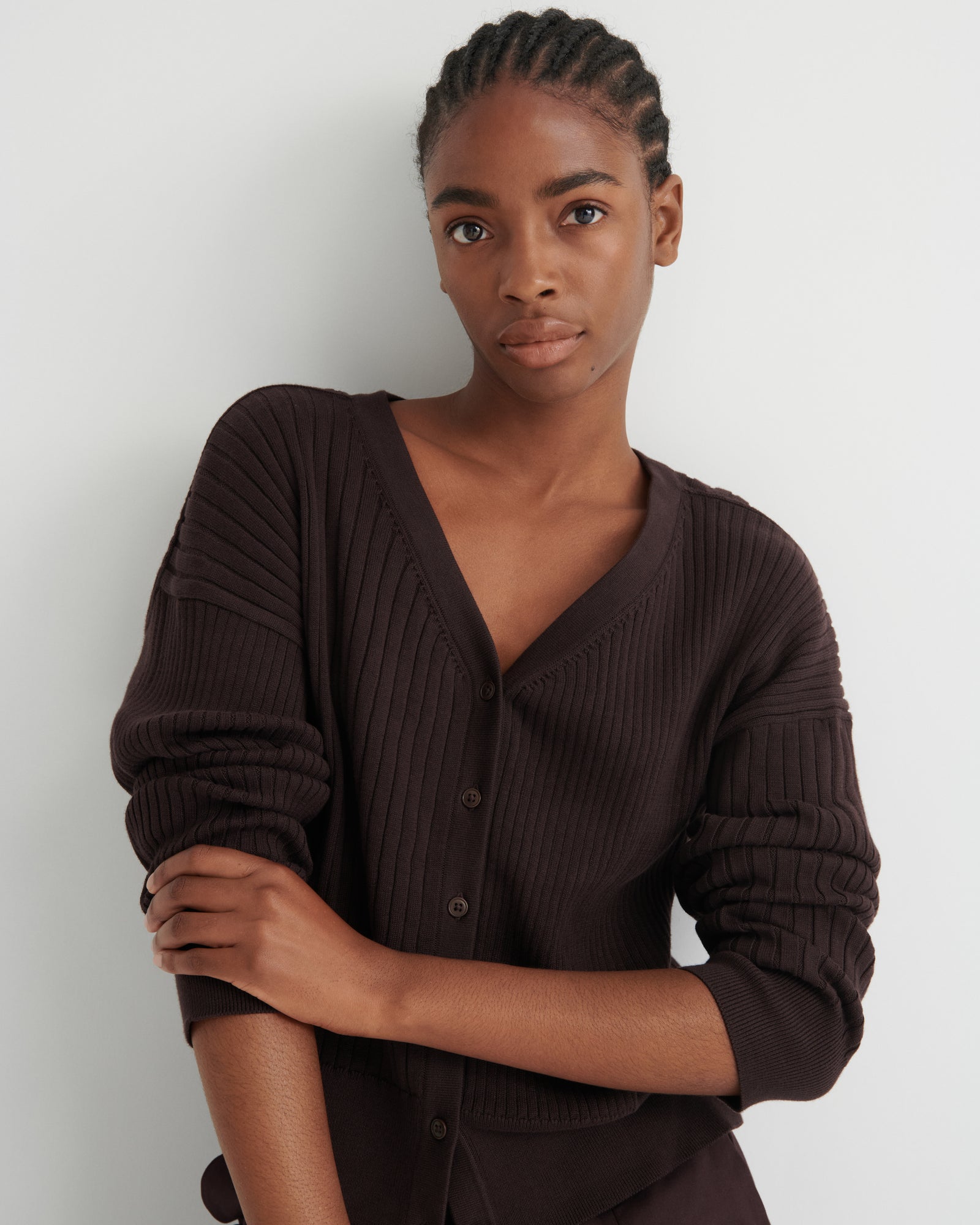 A woman wearing a dark brown ribbed knit cardigan sweater with a V-neck and button-front closure, posed against a plain light gray studio background.