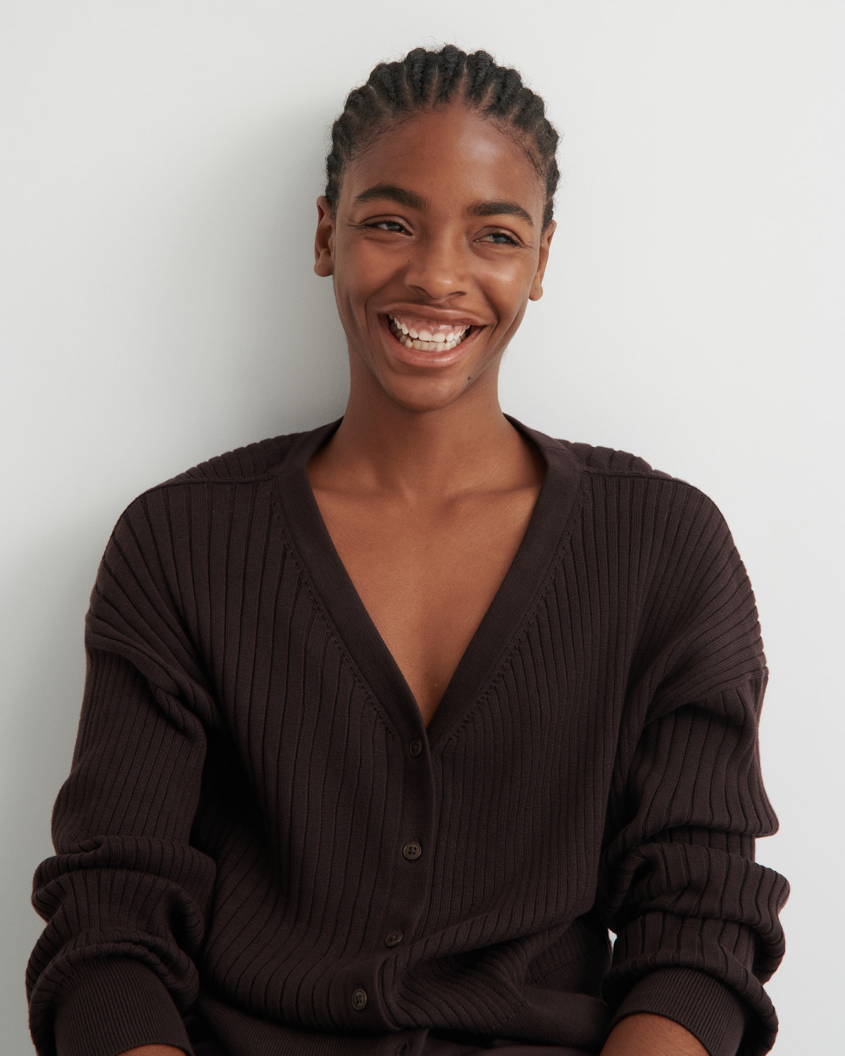 A smiling woman with braided hair wearing a dark brown ribbed cardigan sweater, photographed against a plain light gray background.
