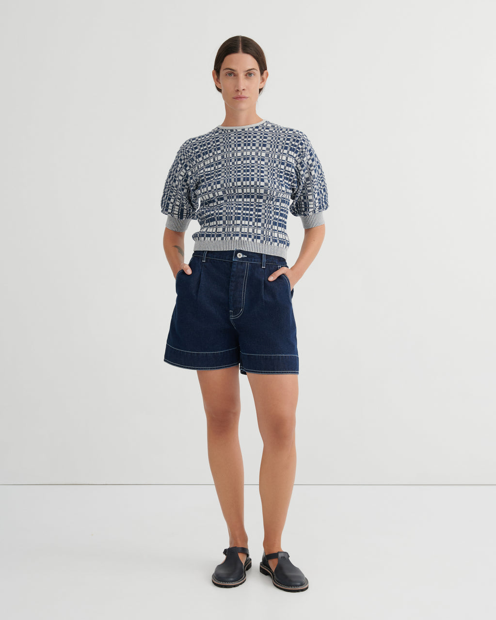 A model stands against a light gray studio background wearing a short-sleeve knit top with a blue and white geometric pattern, dark blue denim shorts, and dark slip-on shoes.