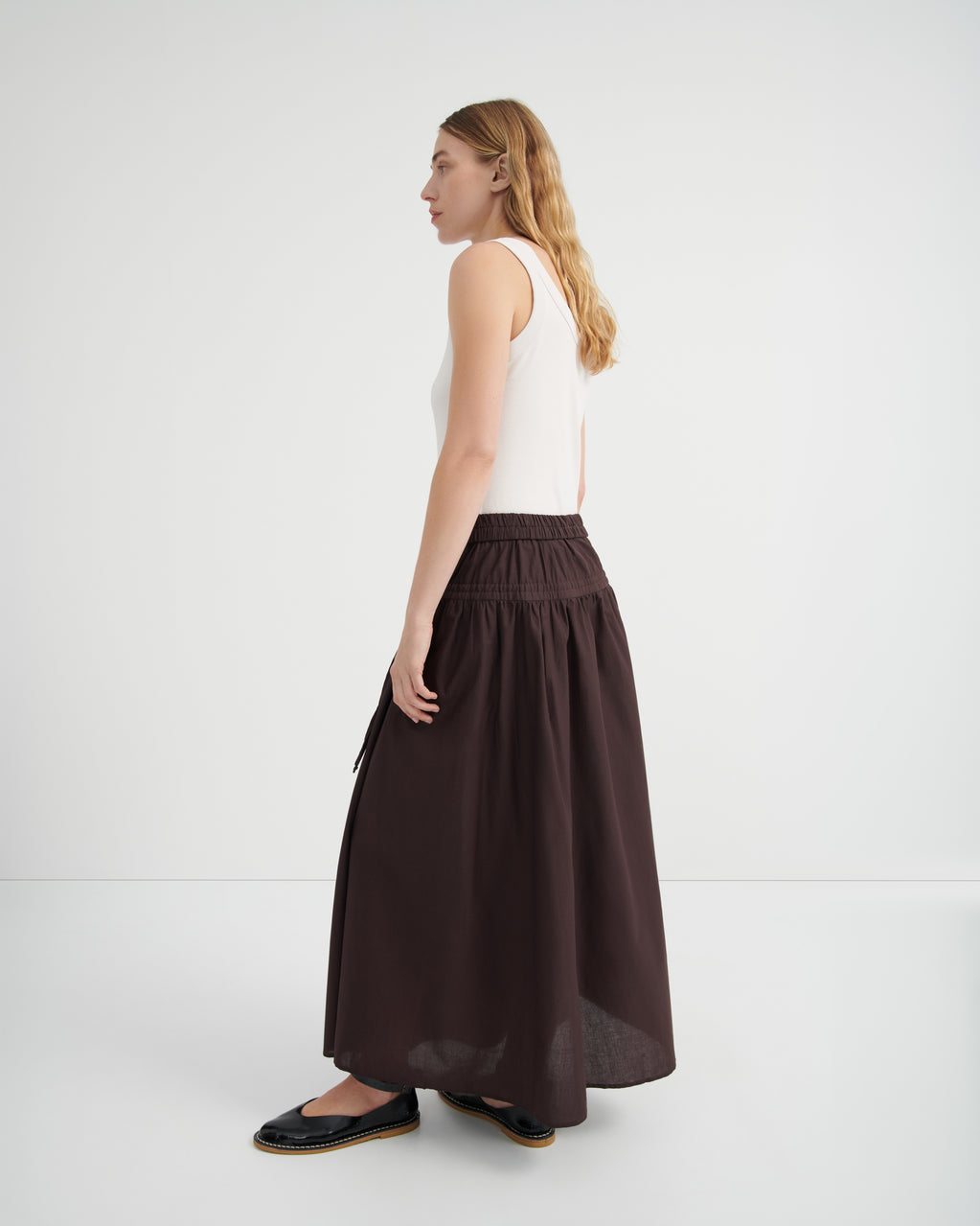 A woman shown from the side profile wearing a white sleeveless top and a long, dark brown gathered skirt.