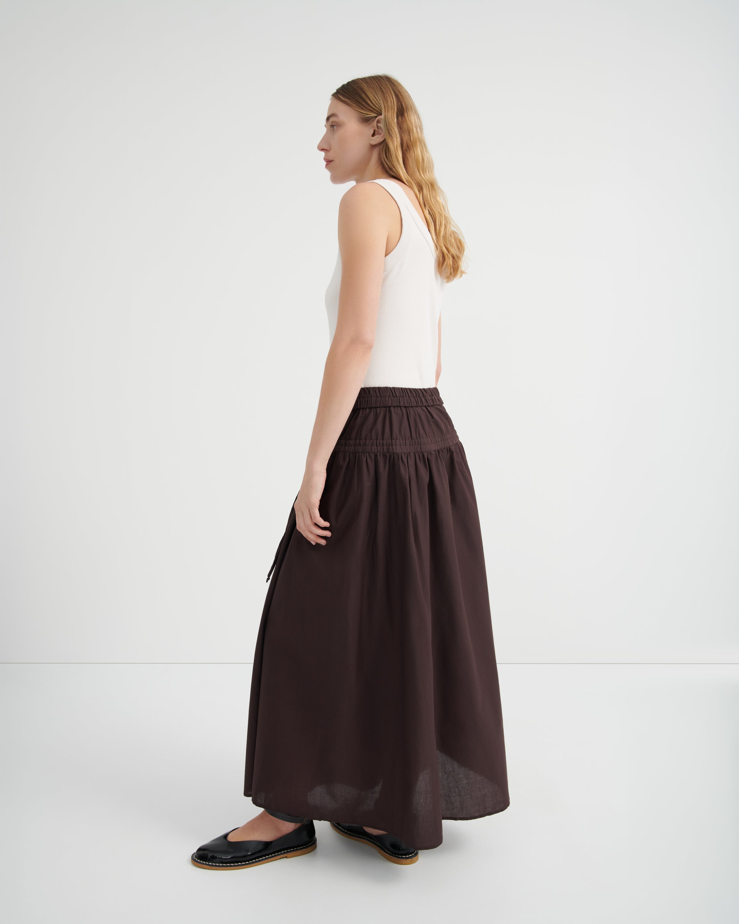 A woman shown from the side profile wearing a white sleeveless top and a long, dark brown gathered skirt.