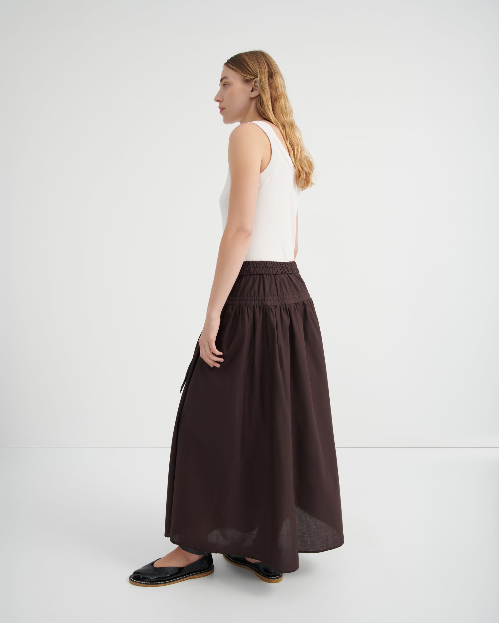 A woman shown from the side profile wearing a white sleeveless top and a long, dark brown gathered skirt.