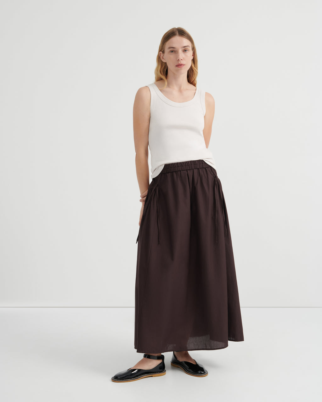 A woman modeling a dark brown high-waisted ruched maxi skirt paired with a white sleeveless top and black flat shoes.
