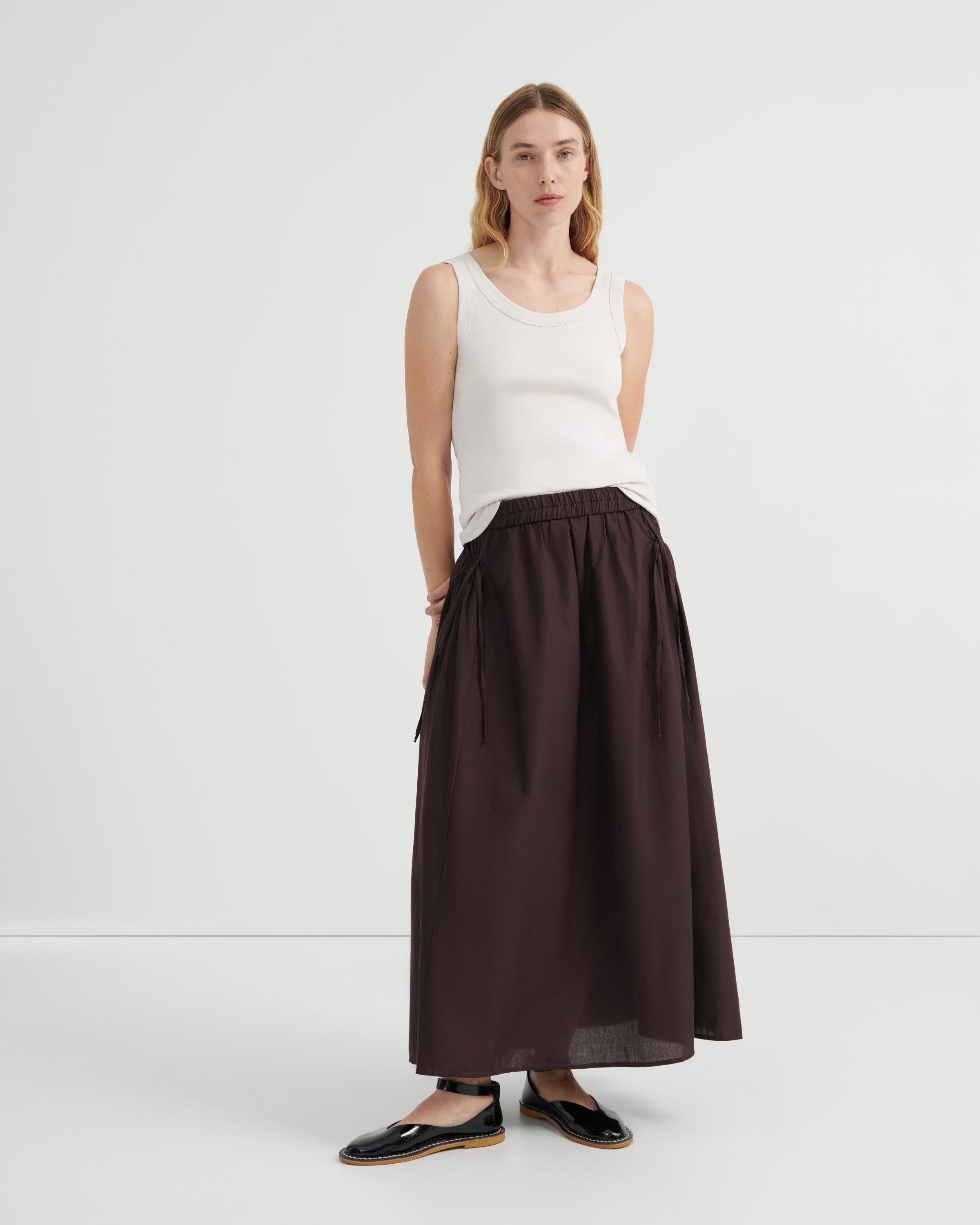A woman modeling a dark brown high-waisted ruched maxi skirt paired with a white sleeveless top and black flat shoes.