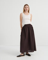 A woman modeling a dark brown high-waisted ruched maxi skirt paired with a white sleeveless top and black flat shoes.