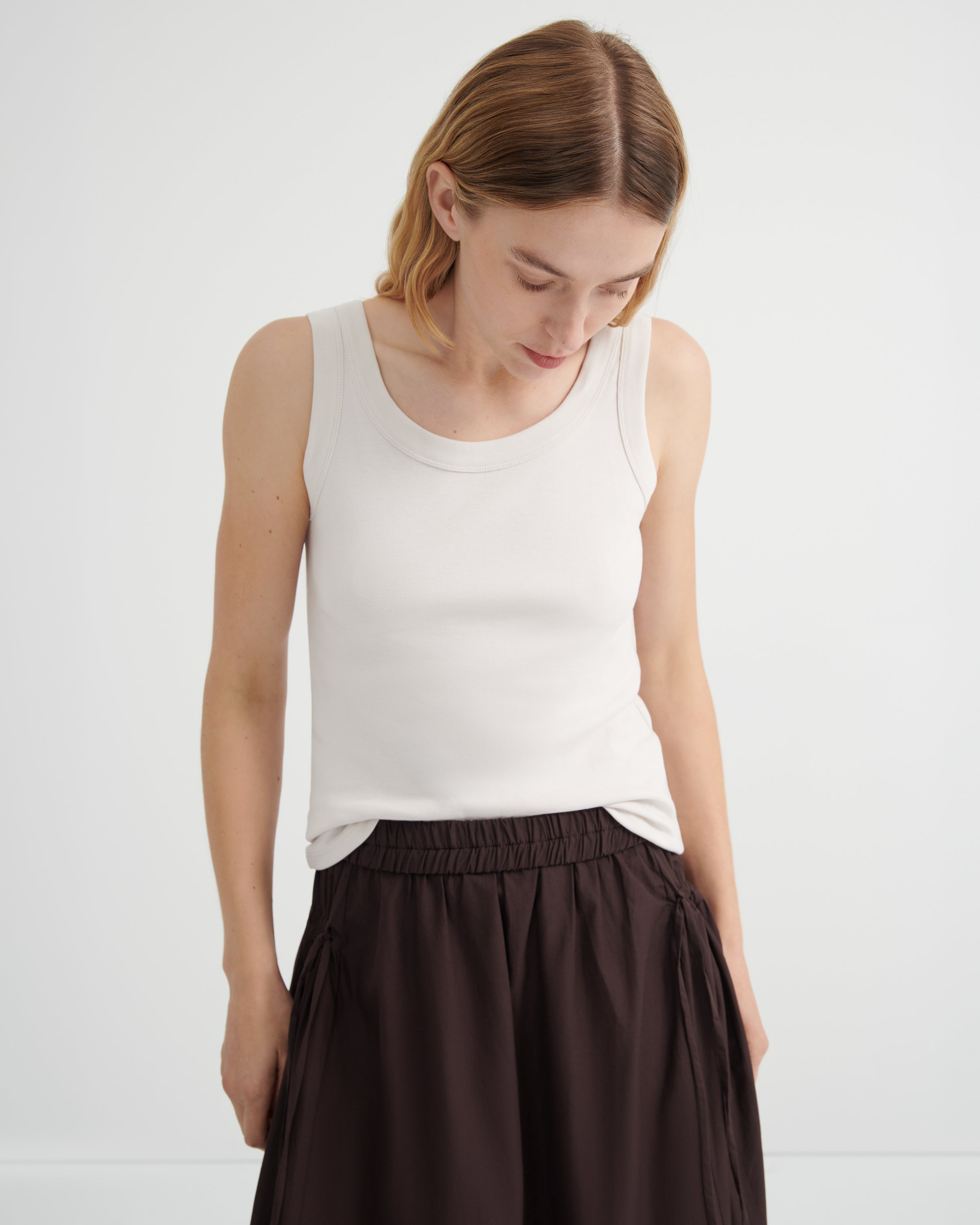A woman wearing a fitted white ribbed tank top and dark brown pleated skirt, looking down.