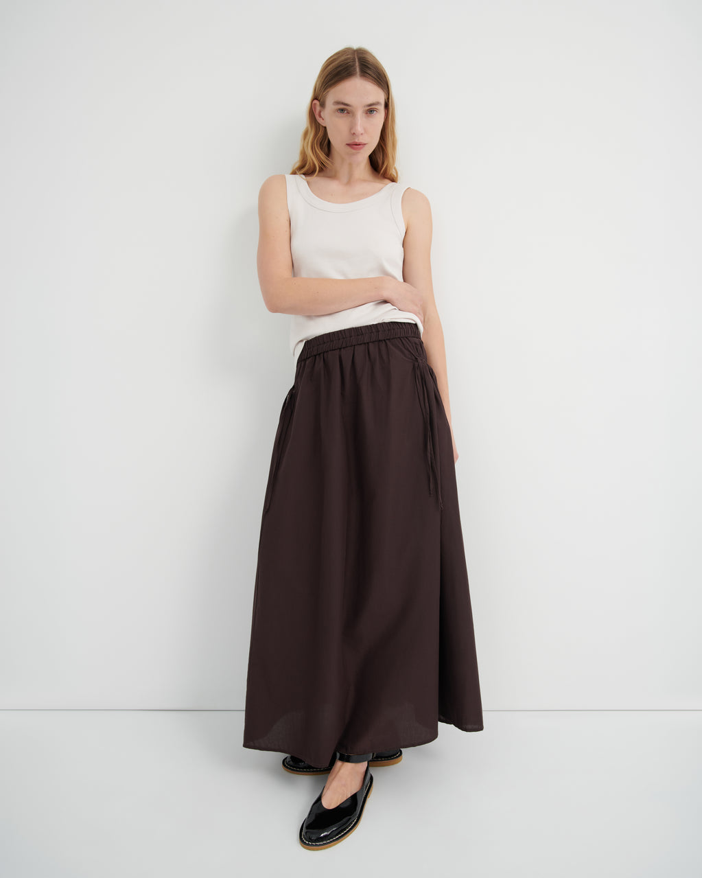 A model wearing a sleeveless white top and a long dark brown skirt with a tie waist, paired with black flats.