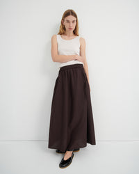 A model wearing a sleeveless white top and a long dark brown skirt with a tie waist, paired with black flats.