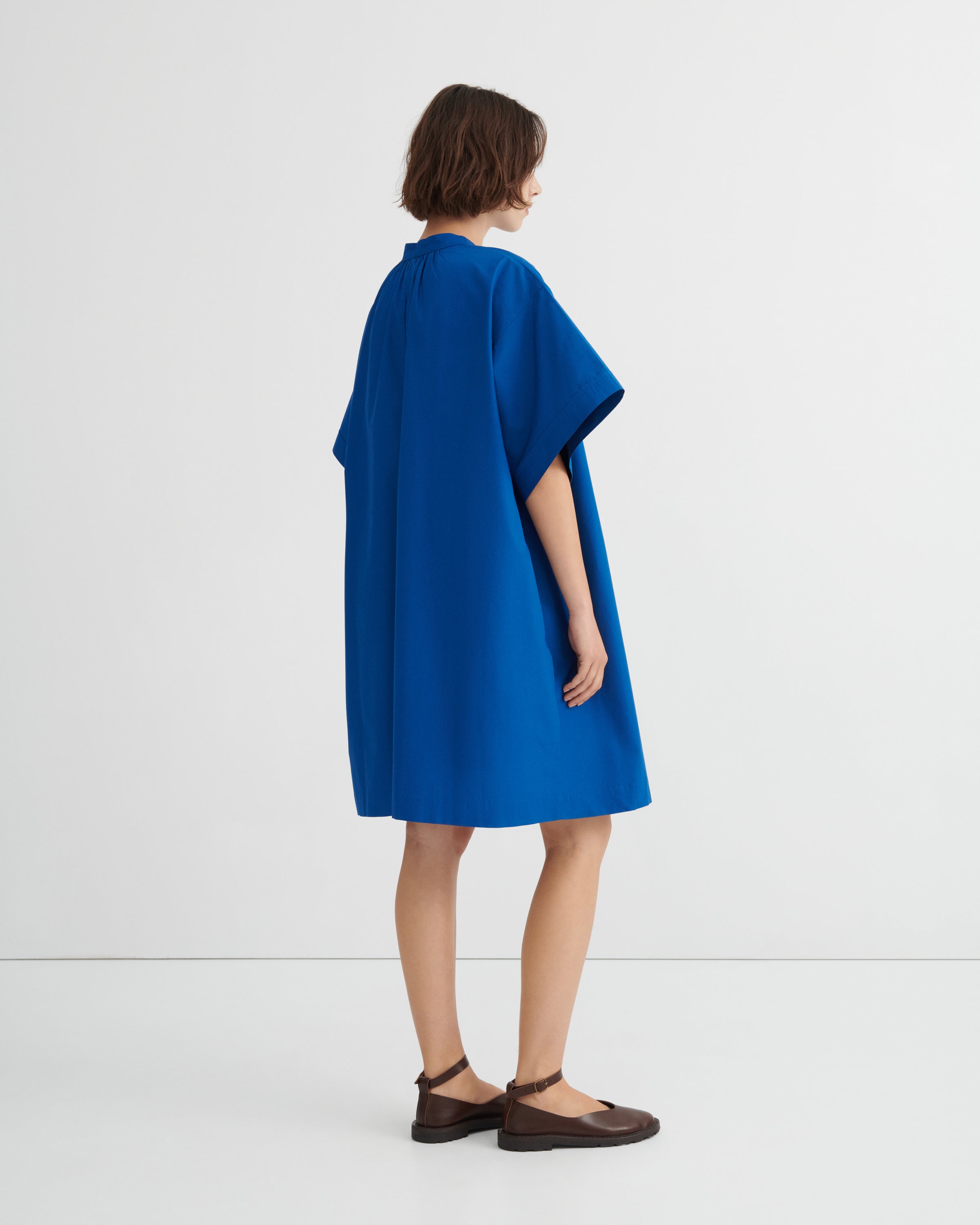 A model shown from the back wearing a bright blue short-sleeved dress with a relaxed silhouette, paired with dark brown flat shoes.