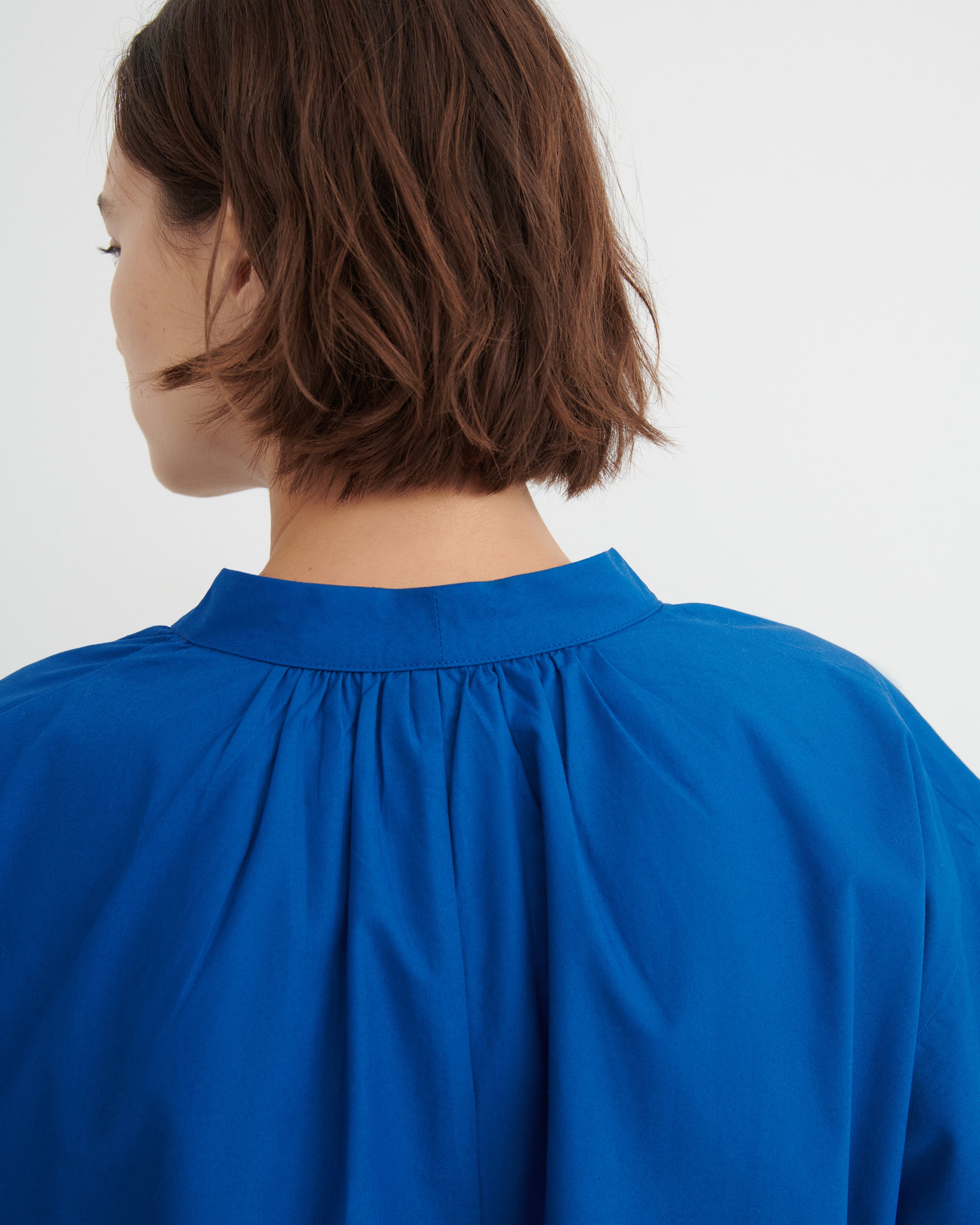 A close-up back view of a person with short brown hair wearing a bright royal blue dress with a gathered, pleated neckline.