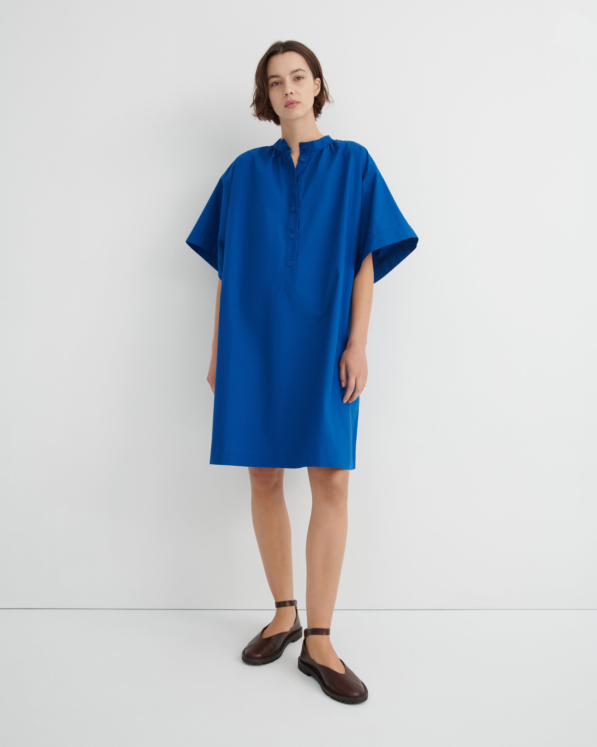 A woman modeling a loose-fitting, short-sleeved blue dress with a buttoned mock neck.