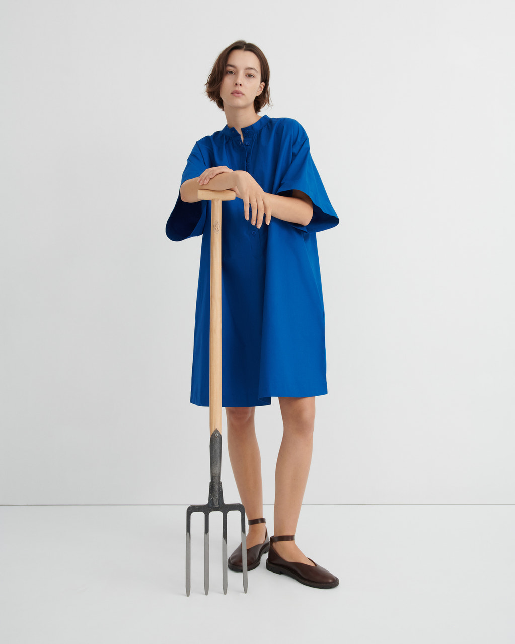 A model wearing a loose, short blue dress and dark brown shoes, holding a large pitchfork with a wooden handle.