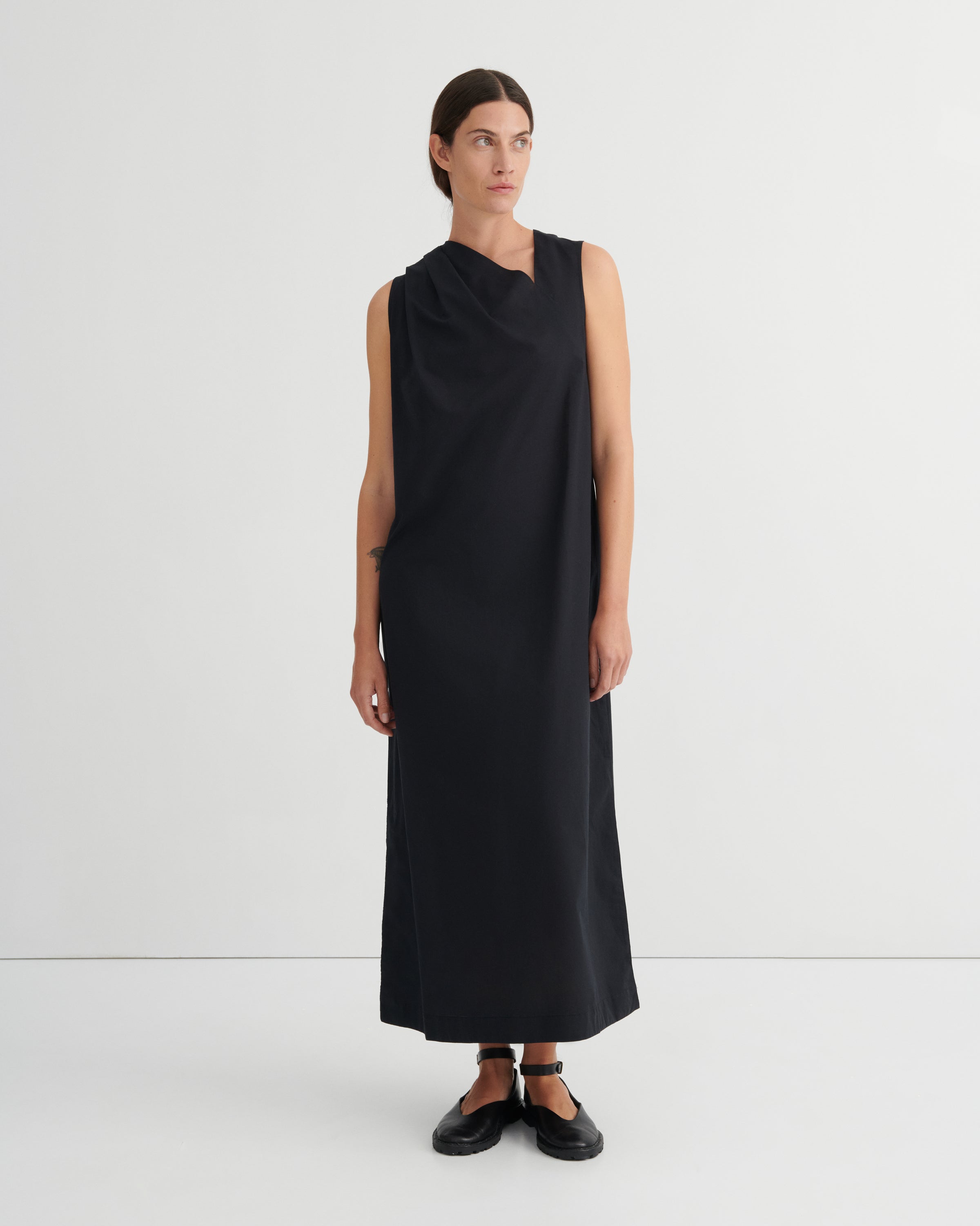 A model wearing a sleeveless, black draped midi dress with a high neckline, paired with black flat shoes, standing against a plain white studio background.