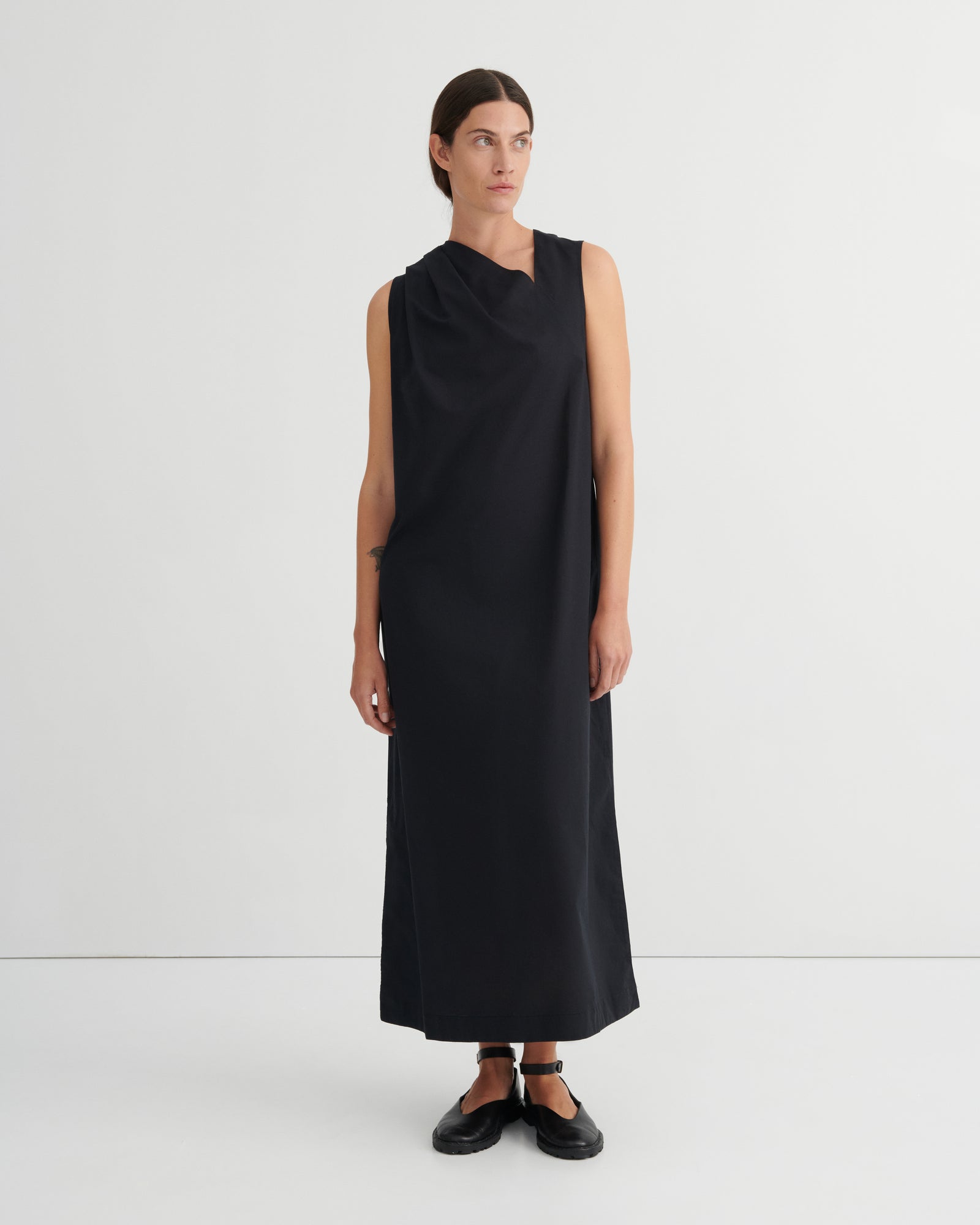 A model wearing a sleeveless, black draped midi dress with a high neckline, paired with black flat shoes, standing against a plain white studio background.