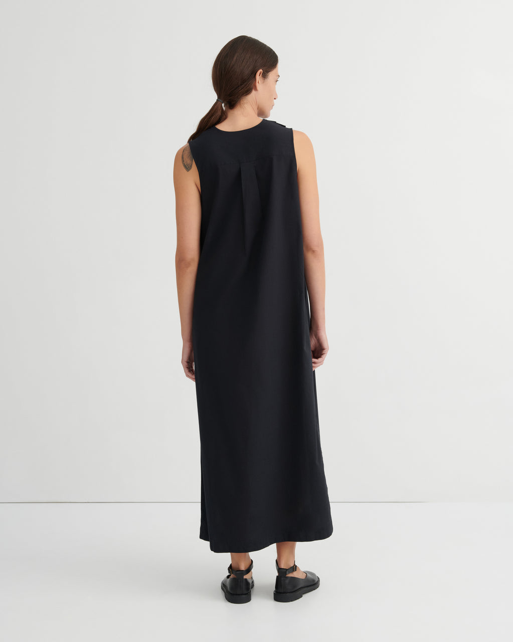 A woman viewed from the back wearing a sleeveless black maxi dress and black shoes, standing against a plain light gray studio background.
