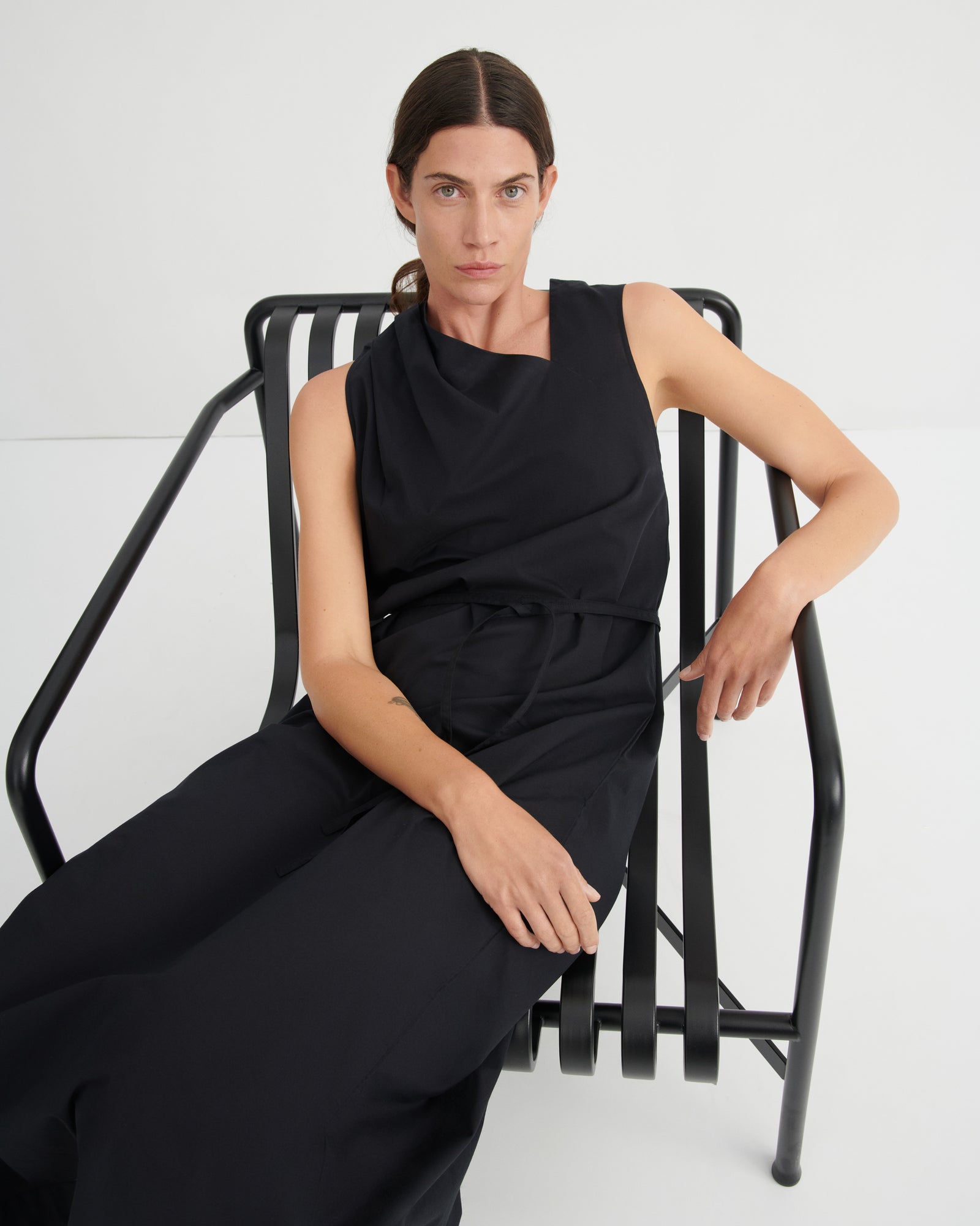 A woman wearing a black sleeveless draped jumpsuit sits on a black metal chair against a clean white studio background.