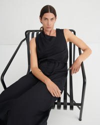 A woman wearing a black sleeveless draped jumpsuit sits on a black metal chair against a clean white studio background.