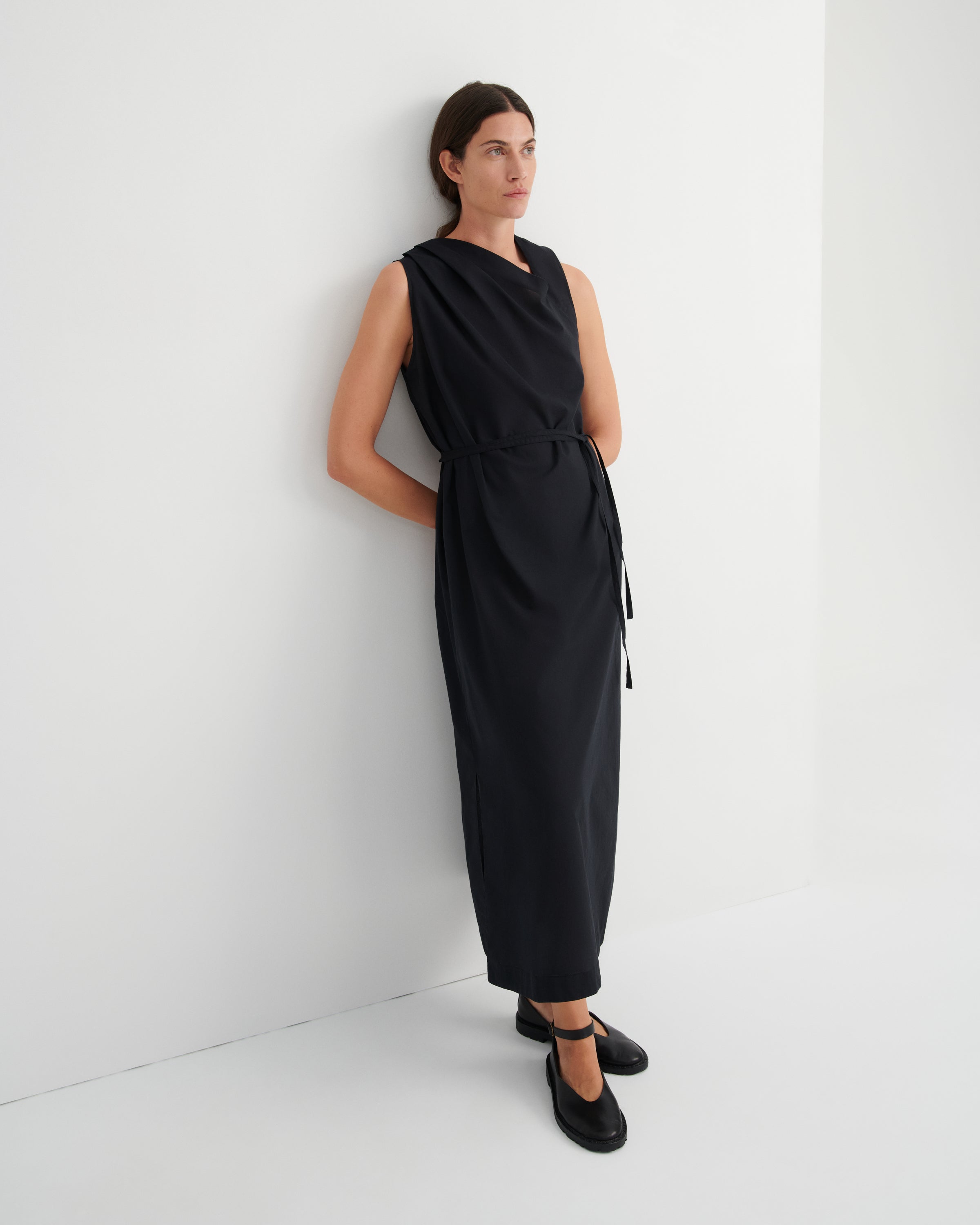 A woman wearing a sleeveless black midi dress with a tie waist and black Mary Jane shoes, standing against a clean white wall.