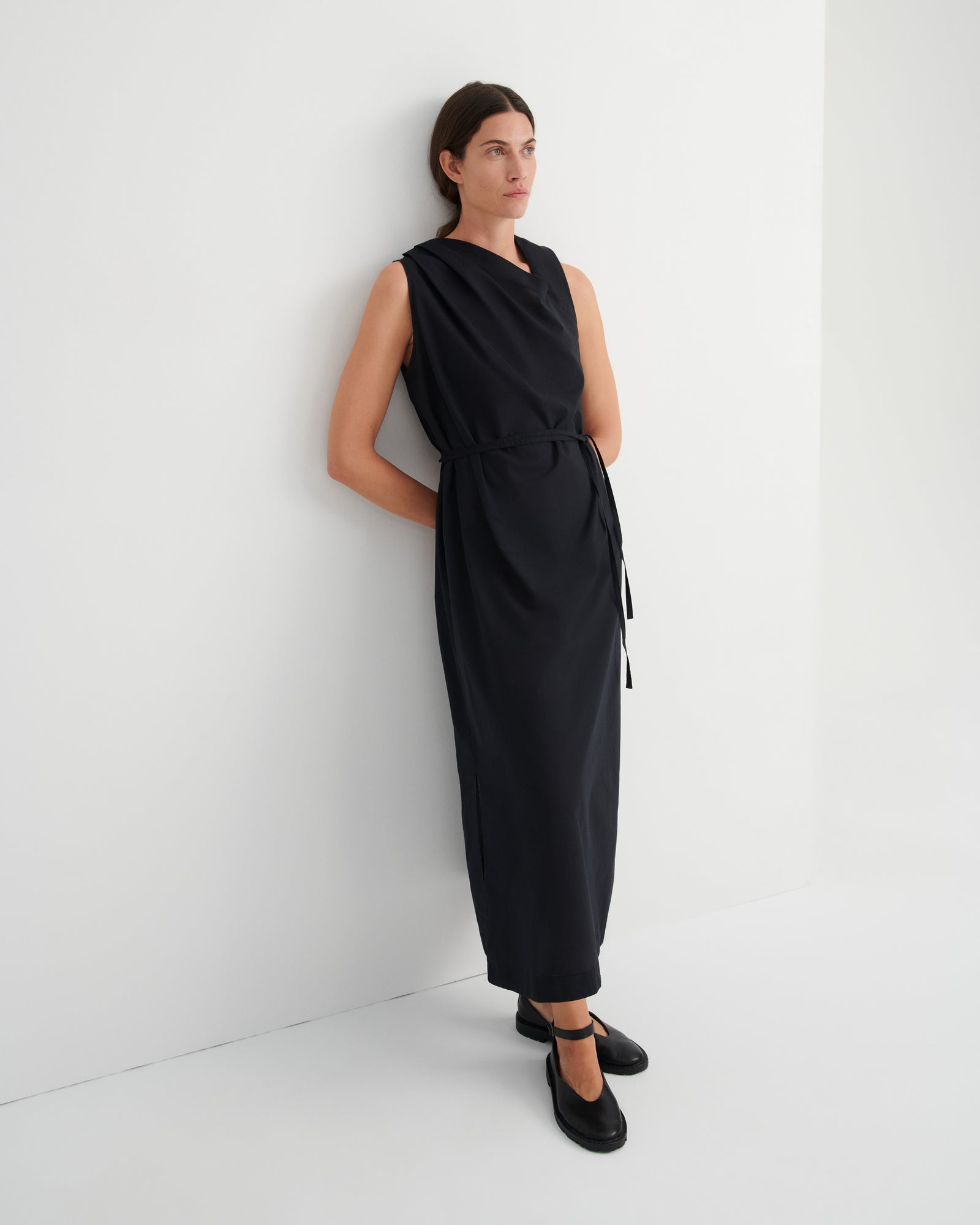 A woman wearing a sleeveless black midi dress with a tie waist and black Mary Jane shoes, standing against a clean white wall.