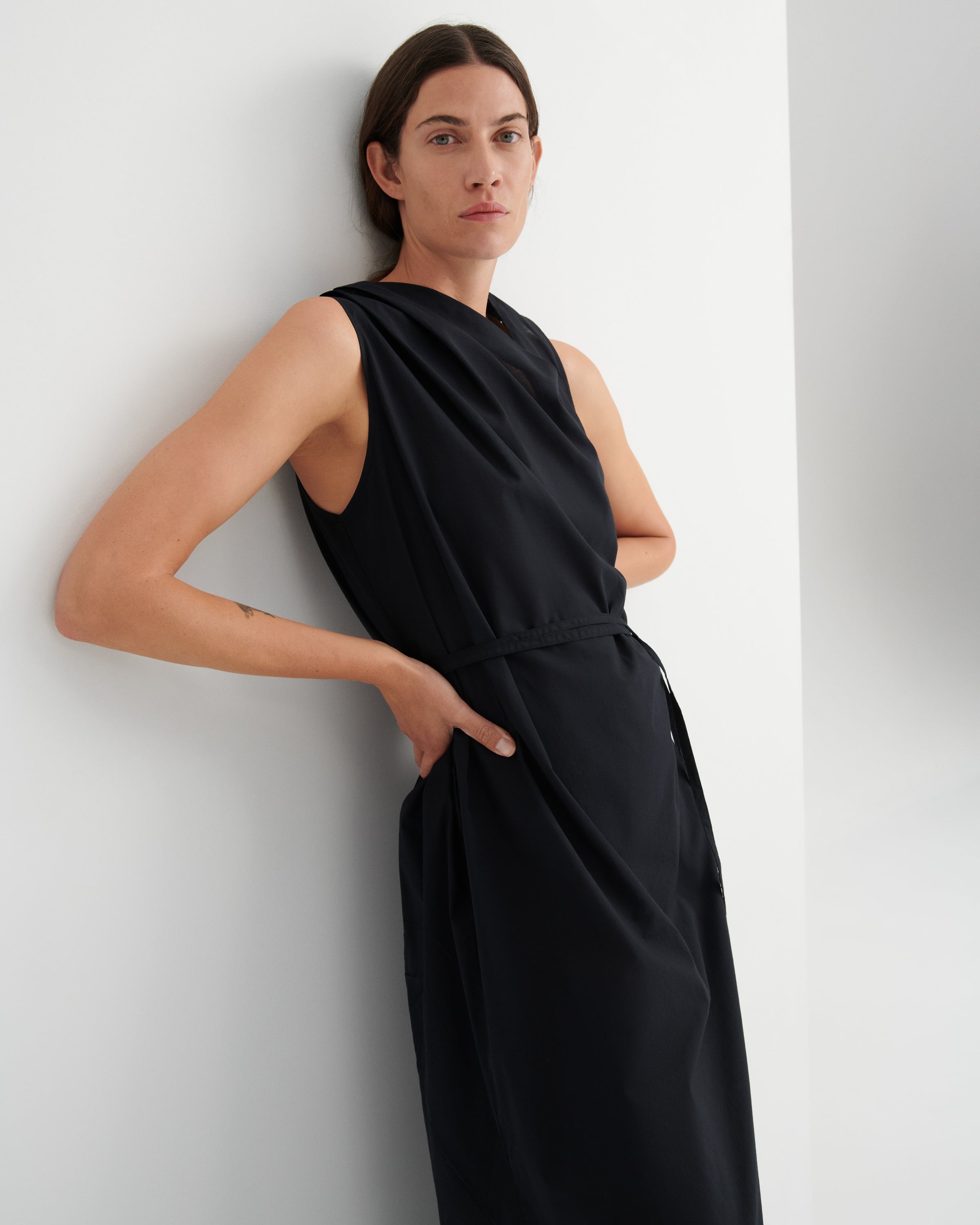 A woman wearing a sleeveless black midi dress with a matching waist tie, posing against a minimalist white wall.