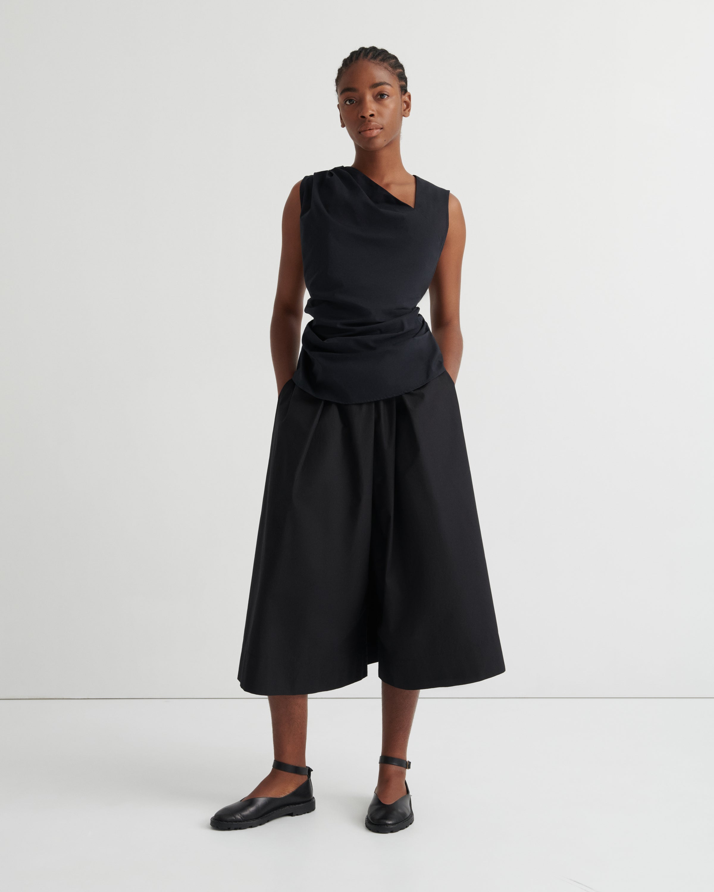 A model stands against a plain light gray studio background wearing a black asymmetrical draped top and matching wide-leg black skirt, paired with black flats.