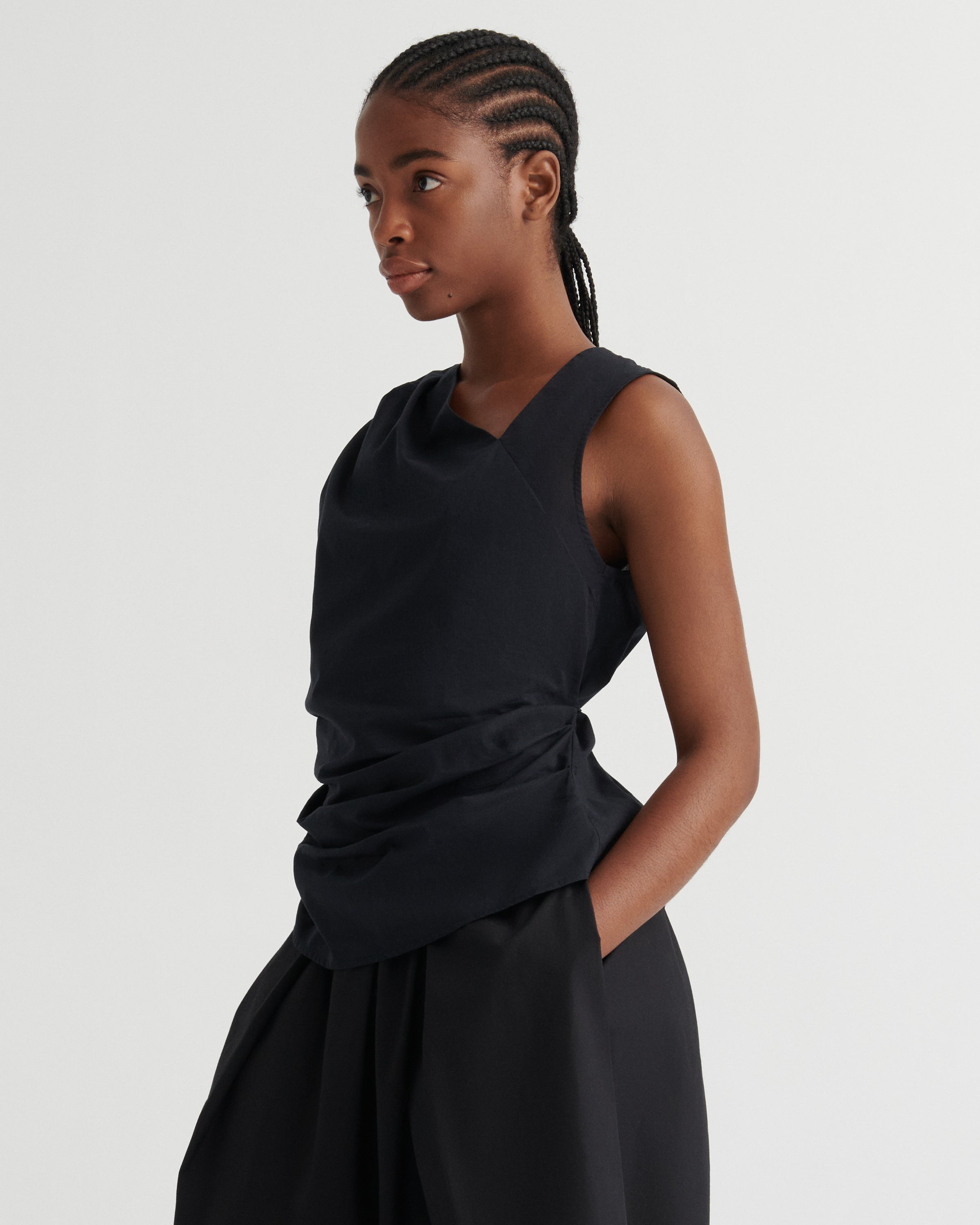A model wearing a black sleeveless wrap-style top and matching black skirt, photographed against a clean light gray studio background.
